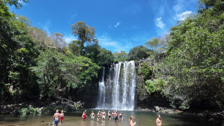 Llanos de Cortés Waterfall falling into its natural pool under a blue sky, with visitors in the water, Guanacaste, Costa Rica