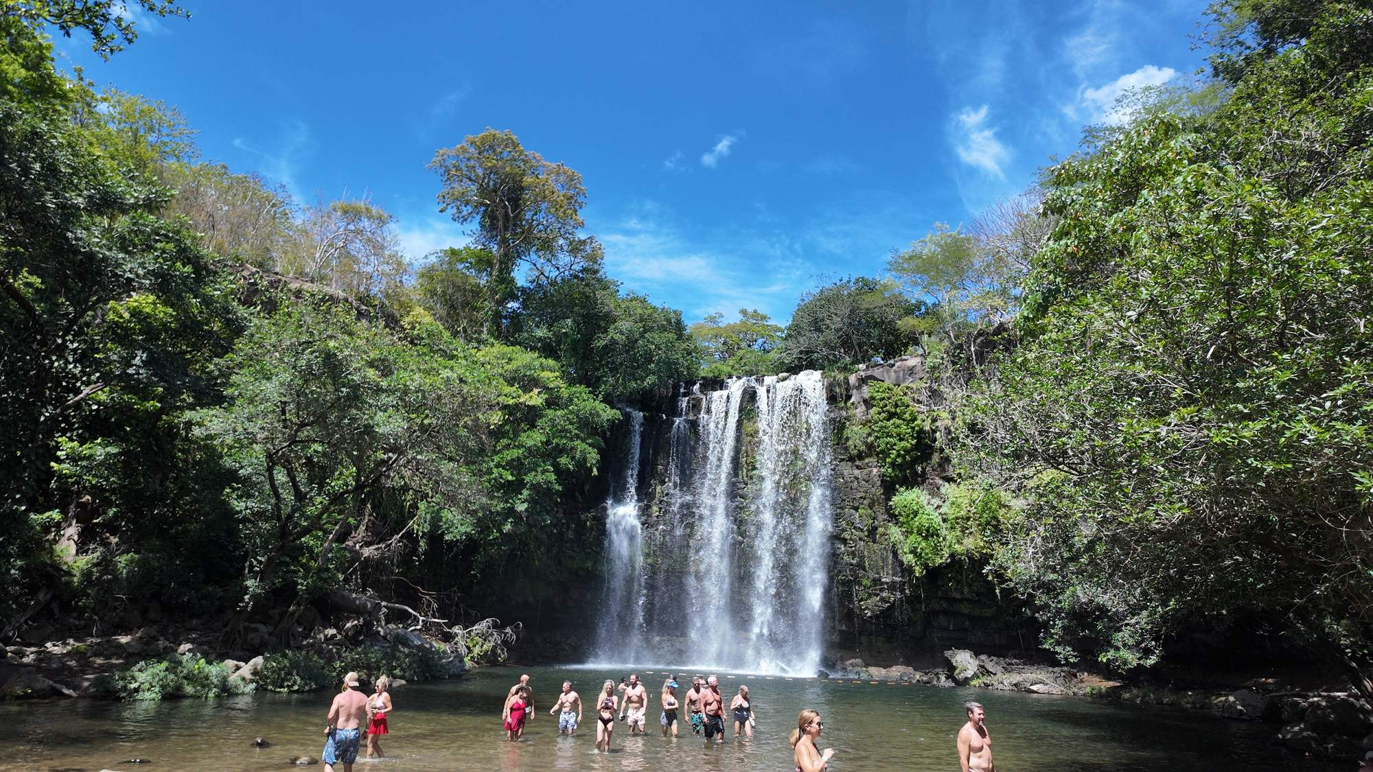 Llanos de Cortés Waterfall falling into its natural pool under a blue sky, with visitors in the water, Guanacaste, Costa Rica