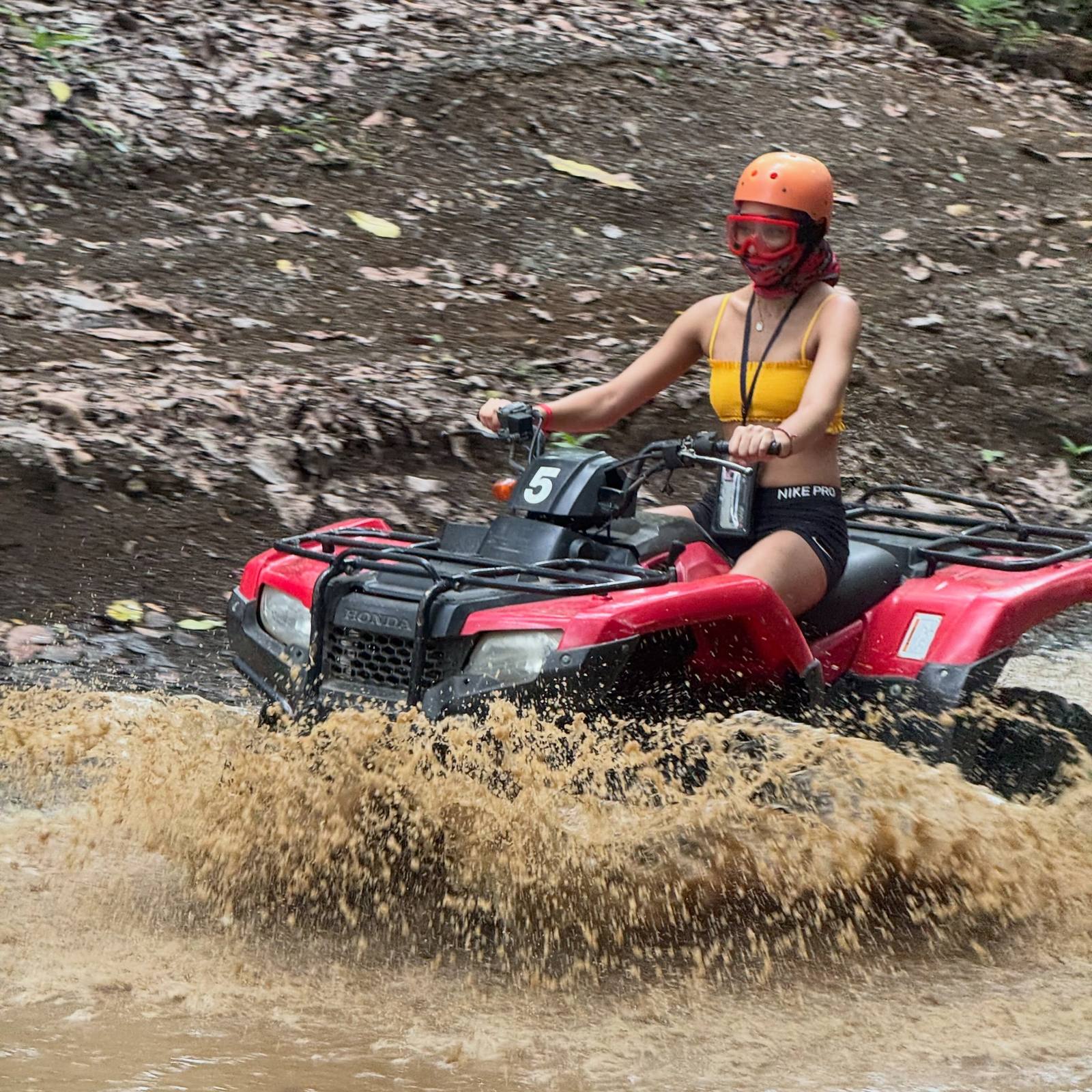 Rider with helmet and goggles driving a red ATV through muddy river water