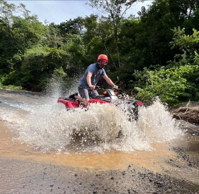 Rider standing on a red ATV while splashing through a river in the forest