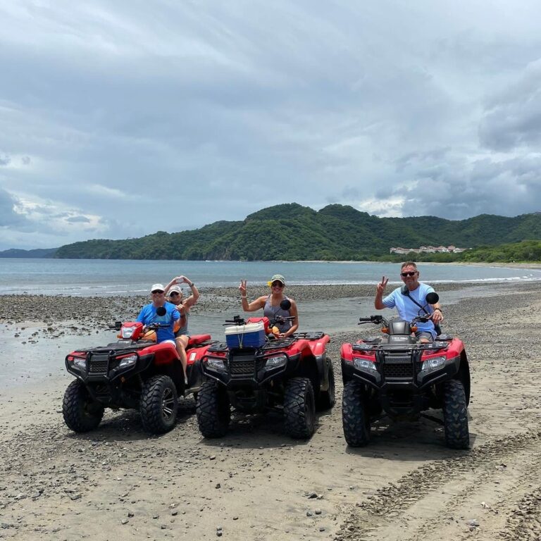 Guests on three red ATVs stopped on a beach with a view of a green bay in Guanacaste