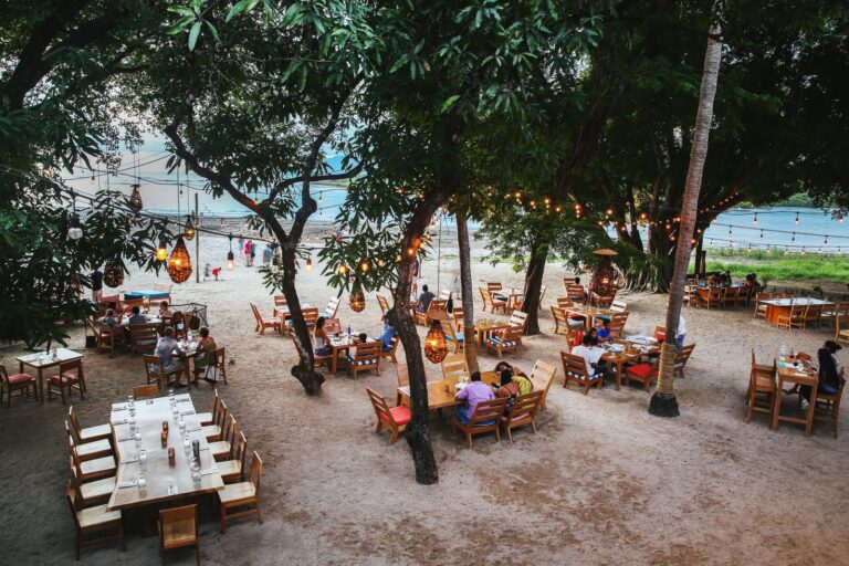 Guests eating at tables on the sand under the trees at a beach restaurant by the ocean
