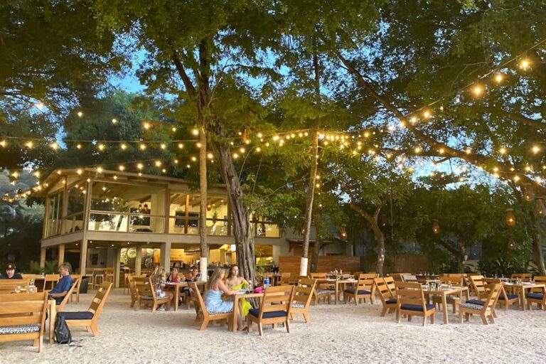 Beachfront restaurant with tables on the sand under trees and string lights in the evening