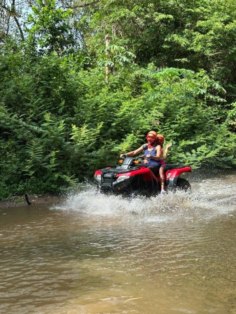 Two riders on a double ATV crossing a river surrounded by green forest