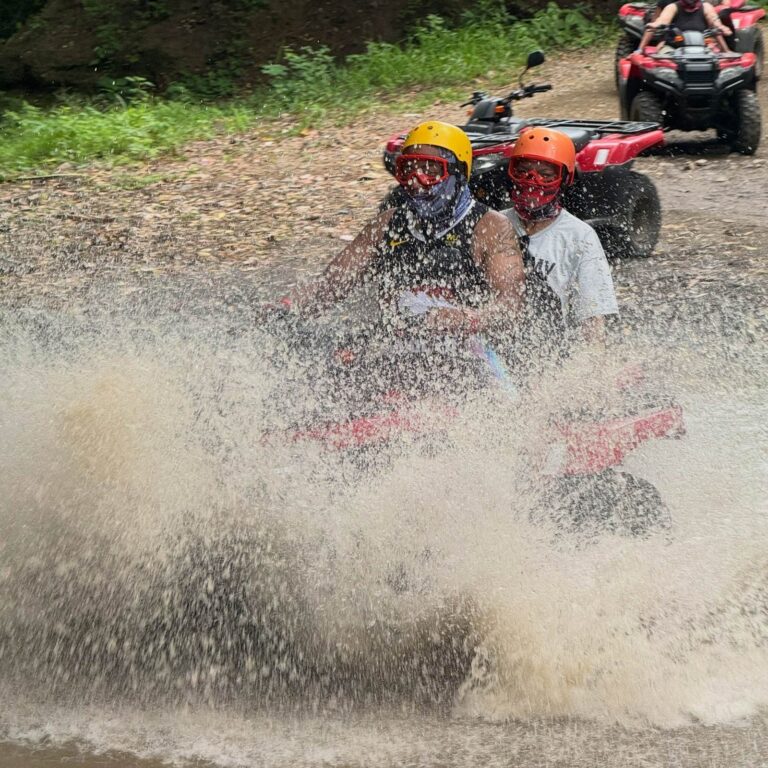 Two riders with helmets and goggles on a double ATV behind a wall of water