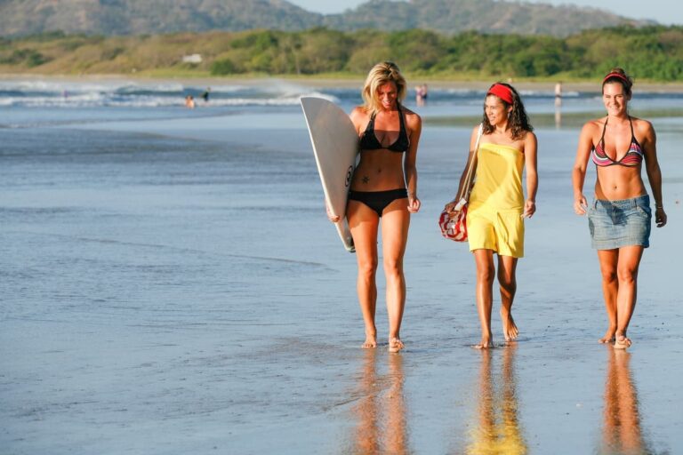 Three friends walking along the beach at low tide, one carrying a surfboard