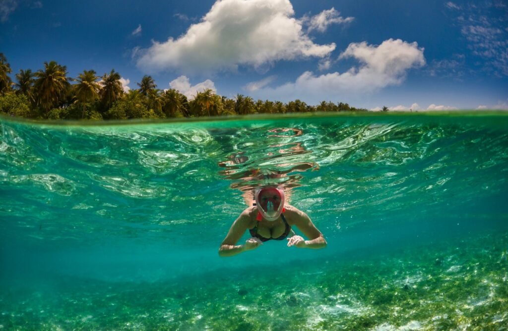 Snorkeler in a full-face mask swimming in clear turquoise water, with palm trees on the shore behind